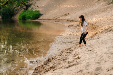 Little girl in white t-shirt, black leggings go down hill closer to river. Kid with long hair run on sand to water.