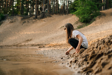 Curious girl with brown hair squatting down on sand near river in forest and going to take grit. Water reflect child.