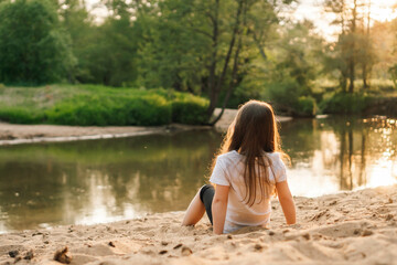Little girl with dark hair sit on sand near forest. Female child in white t-shirt and black shorts have fun in nature.