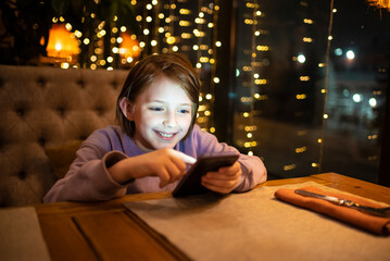 10 year old boy, child plays games on a mobile phone, surfs the Internet using a smartphone, in the evening in a cafe, restaurant, at home