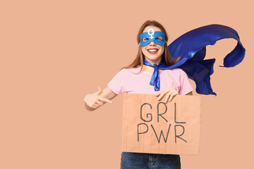 Young woman in superhero costume holding cardboard with text GRL PWR on beige background
