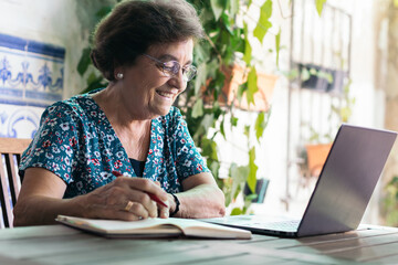 Old woman with glasses using a laptop and a notebook at home - obrazy, fototapety, plakaty
