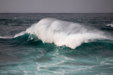 An angry turquoise green colour massive rip curl of a wave as it rolls along a beach. The white mist and froth from the wave are foamy and fluffy. The Atlantic Ocean in the background is deep blue. 