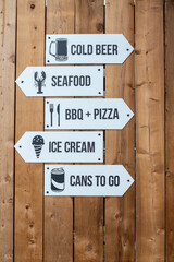 A rustic wooden wall with knotty pine boards. Multiple white arrows with directions and cold beer, seafood, BBQ, pizza, ice cream, and cans to-go signs are stacked in a row. The text is black. 
