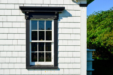 Fototapeta na wymiar The exterior wall of a white cedar shake wooden clapboard covered the house, with a vintage glass double hung window. The antique window has black painted wood trim. There are trees in the background.