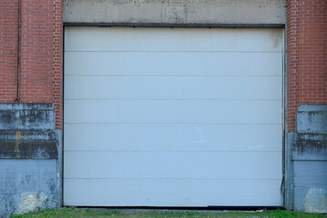A large white metal panel car garage door in a narrow red brick building. The foundation of the building is grey concrete with a black waterproof coating. The ground in front is asphalt. 