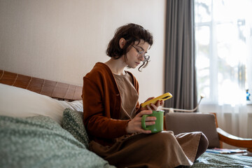 Apathetical teenage girl addicted to smartphone sitting at home in sunny weather, scrolling photos...