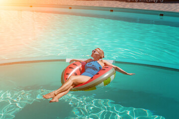 Happy woman in a swimsuit and sunglasses floating on an inflatable ring in the form of a watermelon, in the pool during summer holidays and vacations. Summer concept.