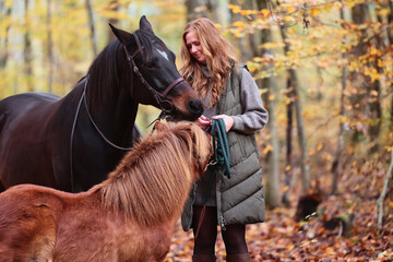 Woman with red hair stands in front of her horses in the autumn forest.