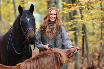 Woman with red hair in a winter coat stands with horses in an autumn forest, head portraits...