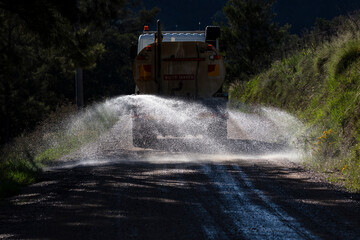 Photograph of a large water tanker spraying water on a dirt road that is under repair running...