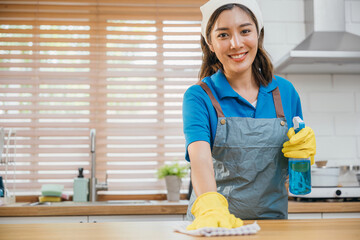 Asian woman in yellow gloves cleans wooden kitchen counter with liquid spray ensuring hygiene....