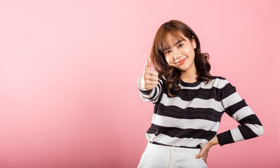 A smiling Asian woman in a studio shot on pink background confidently gives a thumbs-up 'Ok' sign,...