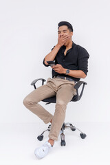 An indolent young man looking at his phone while yawning. Sitting slouched on an office armchair. Laziness in the workplace concept. Isolated on a white background.