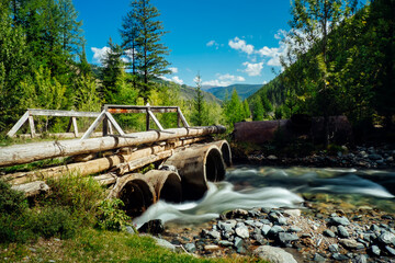 Old wooden bridge across a swift mountain stream in the forest. Water flows quickly through pipes,...