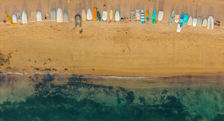 aerial image at a top angle from a drone of the seashore on an Australian beach showing the kayaks and colorful surfboards on the brown sand, and the green sea water and some people walking