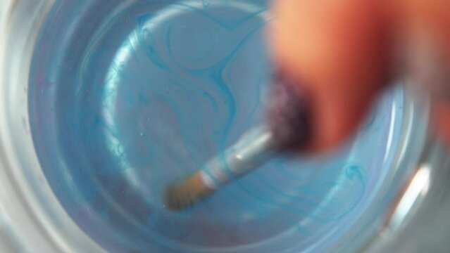 Blue paint disperses from hairs of watercolor paintbrush as it is swirled in clean water cup