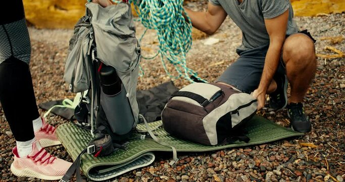 Close-up shot of a girl and a guy disassembling their backpacks and taking out equipment for rock climbing on a rocky beach near the yellow rocks during the day near the sea