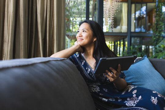 Thoughtful asian woman on couch using tablet at home