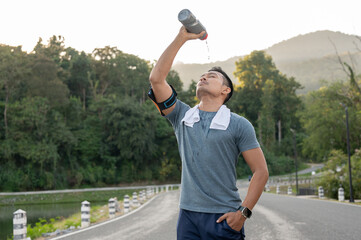 A thirsty, tired Asian man is drinking pouring water from a bottle on his face after a long run.
