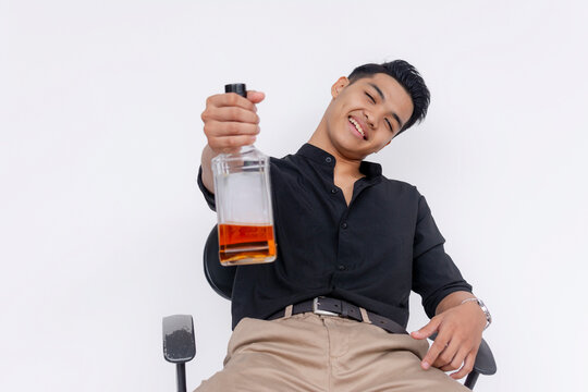 A visible tipsy but friendly young man offers a half-empty bottle of whiskey while slouching on the chair. Isolated on a white backdrop.