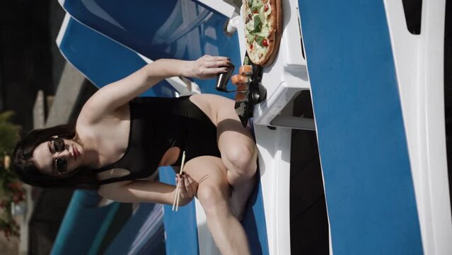 Sexy woman in a black swimsuit eats sushi and pizza near the pool. Hotel holiday concept. Vertical video