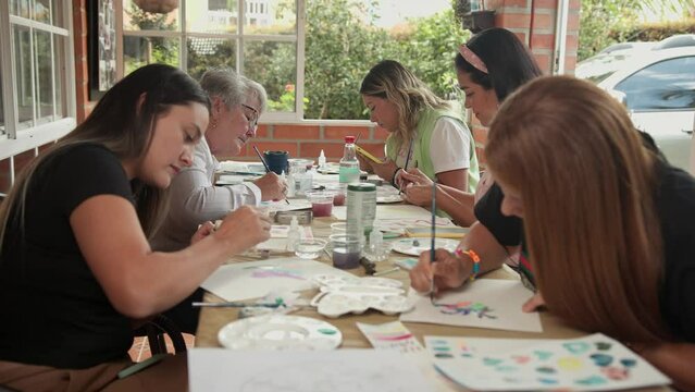 Group of women sit around long table outside paint together with watercolor  in a workshop