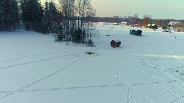 A man submerges himself in a hole cut in the ice of a frozen lake by a sauna - pullback aerial reveal