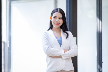 Young happy business asian woman with arms crossed standing int her office and looking at camera.