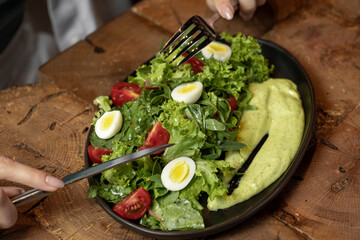 Person eating Fresh green salad with quail eggs, cherry tomatoes and guacamole in black plate on grey background