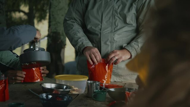 Diverse hiking buddies have meal and rest in gazebo after walking trek. Caucasian hiker cooks freeze dried food in doy pack. Group of tourists during trip or expedition in mountains. Active leisure.