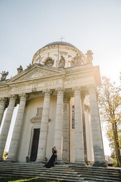 Female kissing male, walk near Baroque Roman Catholic church in Pidhirtsi, Lviv Ukraine. Couple stand on stairs near large columns of ancient temple at sunset. Woman hug man in nature autumn day.