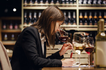 Woman sommelier smelling wine writing notes to tablet using pen sitting in wine cellar among...
