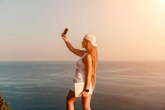 Selfie woman sea. The picture depicts a woman in a cap and tank top, taking a selfie shot with her mobile phone, showcasing her happy and carefree vacation mood against the beautiful sea background