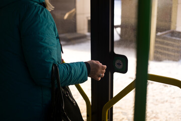 Elderly woman in a warm jacket with a bag in her hand reaches for the button to open the glass doors from inside a modern city bus