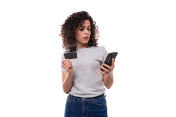 Fototapeta premium young charming brunette curly lady in a gray basic t-shirt holding a mockup of a plastic card and a smartphone on a white background with copy space
