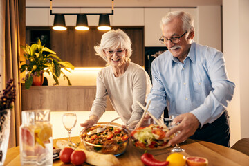 A happy senior couple is putting food on lunch table at home.
