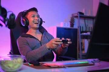 Joyful young Asian man streamer using joystick for playing video game on computer at home.