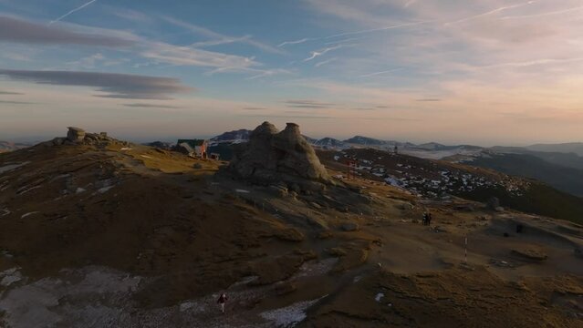 aerial orbit view of bucegi sphinx with tourists next to it on top of mountain during sunset