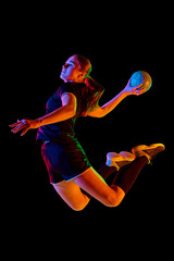 Action shot of young woman, professional handball player passionately playing handball, executing powerful throw against black background in neon light. Concept of sport, energy, dynamic, championship