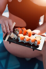 A young woman in a black swimsuit eats a Philadelphia roll near the pool. Concept of rolls and sushi. Relaxation near the pool. Summer season