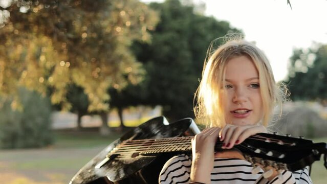 a hipster girl of light appearance with a piercing in her nose stands in the park holding a black guitar on her shoulder, the video was shot around a teenager. High quality FullHD footage
