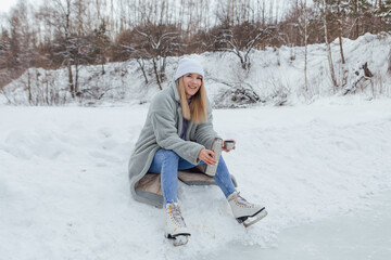 Lovely young woman relaxing after riding ice skates and drinking hot drink from thermo pot on the ice rink.