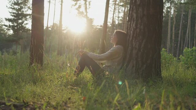 Lady leans on tree trunk bark in forest at sunset. Young woman rests in fresh air thinking about mental health improvement. Coping with personal problems in nature