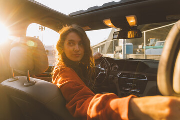 Young pretty woman in a car in sunshine light, behind the wheel, looking back. - obrazy, fototapety, plakaty