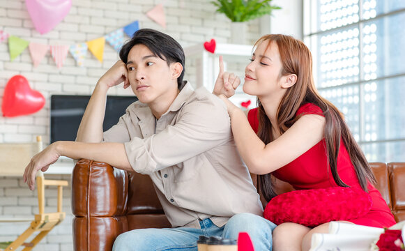 Asian young beautiful female girlfriend sitting cuddling smiling on cozy sofa in decorated living room showing little finger reconciling handsome upset sulking male boyfriend at home on valentine day