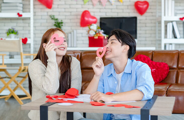 Asian young handsome male boyfriend sitting on sofa smiling cuddling with beautiful female girlfriend holding red heart shape paper cutting in romantic decorated living room celebrating valentine day