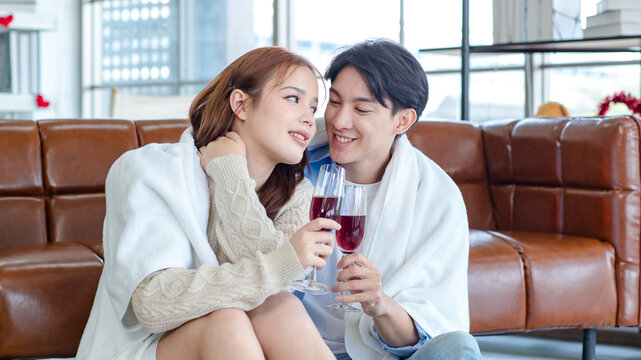 Asian young handsome male boyfriend sitting smiling cuddling together with beautiful female girlfriend holding tall wine glass cheers toasting in romantic decor living room celebrating valentine day