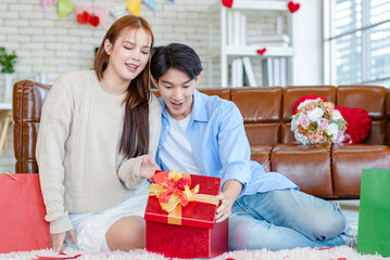 Asian young beautiful female girlfriend sitting smiling opening unboxing red present gift wrapped box on cozy sofa from handsome male boyfriend in romantic decorated living room on valentine day