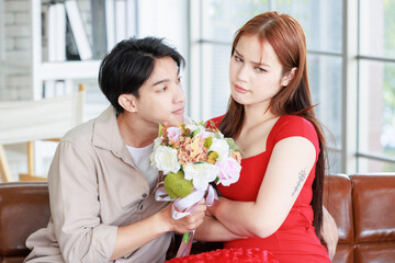 Millennial Asian young handsome male boyfriend reconciling beautiful female sulking upset angry girlfriend in red dress with white roses flower bouquet sit crossed arms on sofa in living room at home